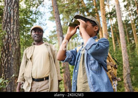 Cute youthful boy looking far away through binoculars while hiking in pine tree forest with his grandfather on summer weekend Stock Photo