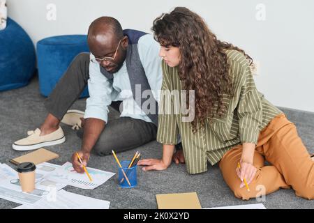 Two young intercultural economists in casualwear discussing papers with financial graphic data while sitting on the floor in office Stock Photo