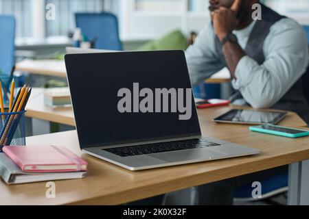 Laptop with black screen on workplace of modern office worker or employee on background of young African American manager Stock Photo