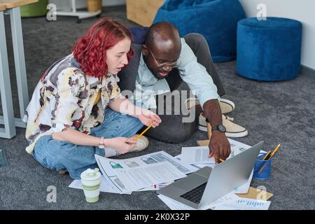 Two young intercultural coworkers discussing data on laptop screen while African American male economist pointing at electronic document Stock Photo