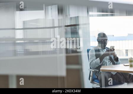 Young serious African American businessman in formalwear scrolling in smartphone while sitting by workplace in office with transparent walls Stock Photo