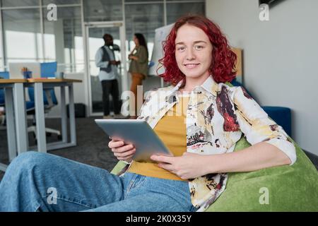 Happy young businesswoman in casualwear using tablet while sitting in armchair in openspace office against two colleagues Stock Photo