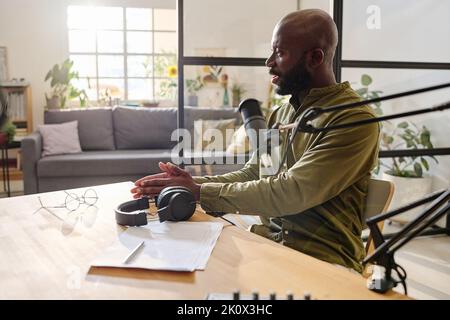 Young confident black man in casualwear explaining something during speculation while sitting by desk and talking in microphone Stock Photo