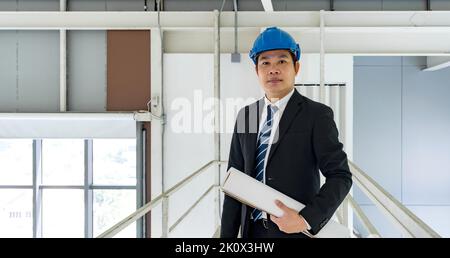 Asian businessman in black suit wearing a blue construction hat,  holding construction drawing. Inspector inspect the orderliness of the factory on th Stock Photo
