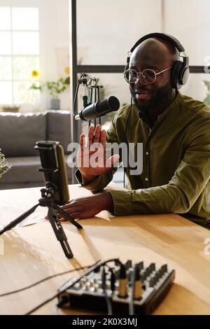 Young male blogger or host in casualwear waving hand to his online audience and looking in smartphone camera during livestream Stock Photo