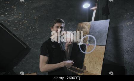 Smiling artist stands next to painting and holds palette with brush in his hands Stock Photo