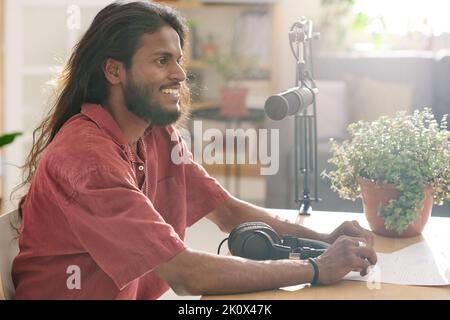 SIde view of young male host sitting in front of microphone by desk and talking to invited guest while recording their conversation Stock Photo