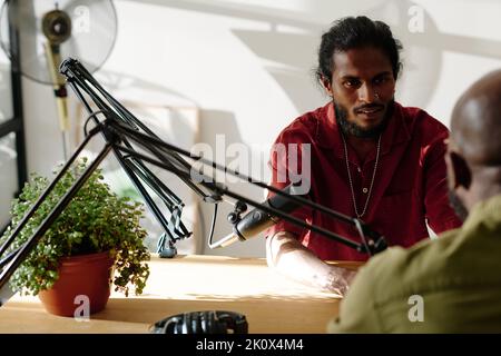 Serious young man sitting in front of microphone by desk and looking at guest invited to studio for recording interview or discussion Stock Photo