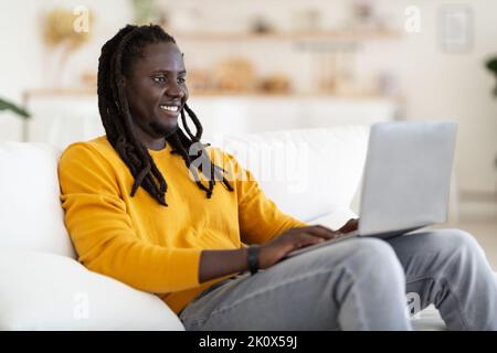 Freelance Concept. Smiling Black Man With Dreadlocks Working With Laptop At Home Stock Photo