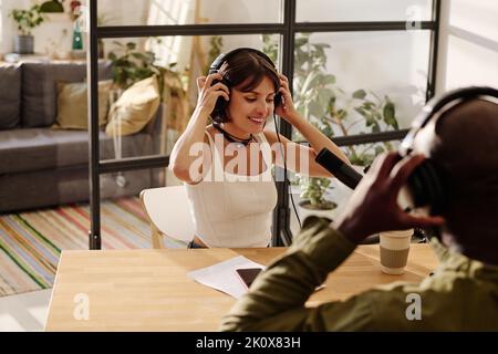 Happy young female host and her African American male guest putting on headphones while sitting by desk in front of one another Stock Photo