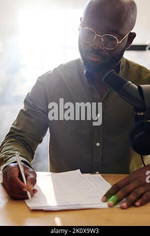 Young bald black man reading questions on paper prepared for interview with guest while sitting by desk in studio in front of microphone Stock Photo