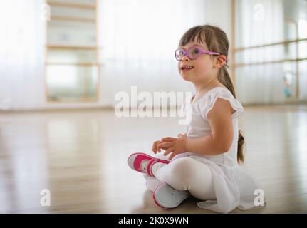 Little girl with down syndrome at ballet class in dance studio,sitting and resting. Concept of integration and education of disabled children. Stock Photo