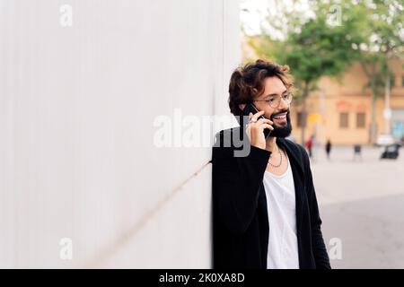 smiling young man with beard leaning in a white wall talking happy on mobile phone, concept of urban lifestyle and technology of communication, copy s Stock Photo