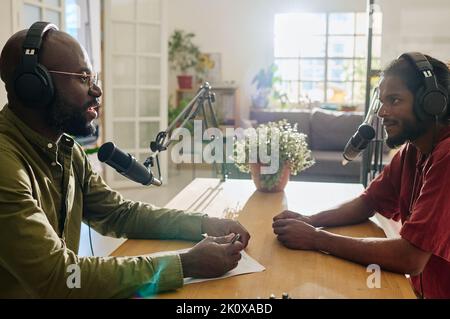 Side view of two young intercultural men sitting by desk in front of one another and talking in microphones during discussion Stock Photo