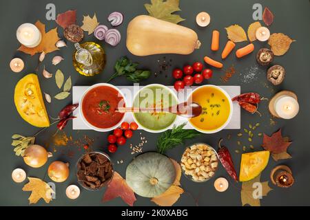 Thanksgiving holiday table with set of different vegetable cream soups with wooden spoon and ingredients, candles and autumn leaves on a dark grey bac Stock Photo