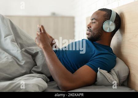 Afrifcan American Man Using Phone Wearing Headphones Lying In Bedroom Stock Photo