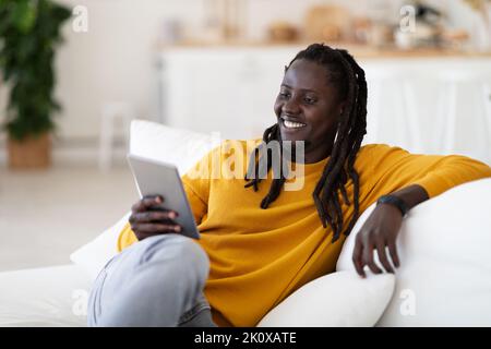 Smiling black man watching videos on digital tablet while resting on couch Stock Photo