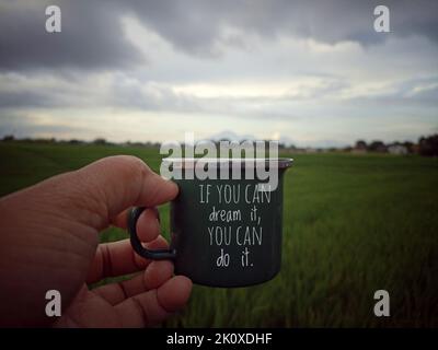 Motivation words on coffee cup. Person holding small green cup in hand on blue gray sky over the green field background with inspirational quote. Stock Photo