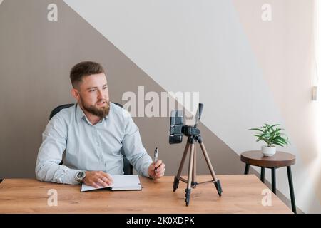Young bearded businessman working in office sitting at table using smartphone for an online meeting making notes in diary. Freelancer works remotely. Student is preparing for seminar. Online marketing Stock Photo