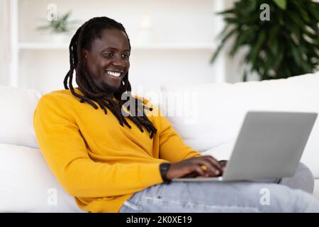 Online Communication. Happy Black Guy Sitting On Couch And Typing On Laptop Stock Photo