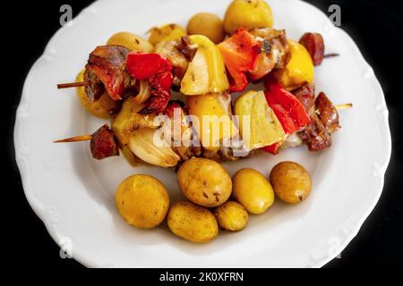 Skewer with pieces of pork meat, red paprika, sausage, bacon and onion and boiled potato in skin on white plate, black background, closeup. Stock Photo