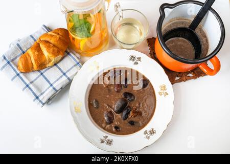 Giant bean soup with dried tomato in plate and in orange pot with ladle, bread on towel, orange drink in pitcher and glass on white background. Stock Photo