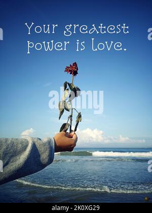 Inspirational quote - Your greatest power is love. With person holding dry red rose in hand on blue summer sky background on the beach. Life strength. Stock Photo