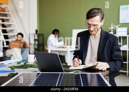 Young businessman making notes in notebook while working at table with laptop and solar panel Stock Photo