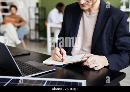 Close-up of businessman writing plans in his notebook while working at table on laptop Stock Photo