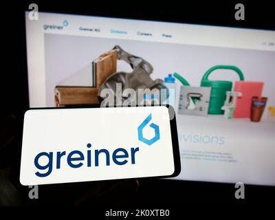 Person holding mobile phone with logo of Austrian plastics company Greiner AG on screen in front of business web page. Focus on phone display. Stock Photo