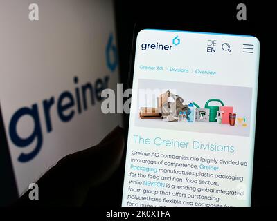 Person holding cellphone with webpage of Austrian plastics company Greiner AG on screen in front of logo. Focus on center of phone display. Stock Photo