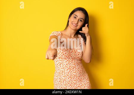 Young beautiful woman wearing summer dress over yellow isolated background smiling doing talking on the telephone gesture and pointing to you. Call me Stock Photo