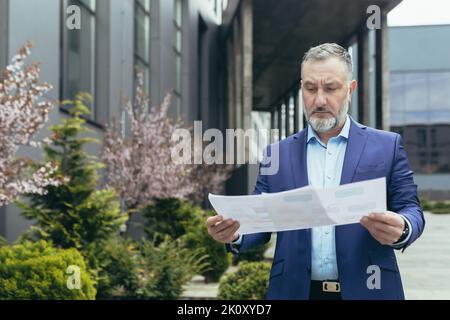 Real estate agent. Handsome senior serious man in suit and beard standing on street near houses, office center, holding documents in hands, plan of apartments. Stock Photo