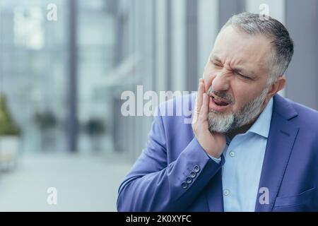 Toothache. Close-up photo. Senior man, businessman, office worker standing on the street near work, holding his cheek, feeling unbearable toothache, grimacing, needs help. Stock Photo