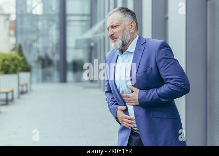 Senior man, businessman, office worker in a business suit stands near the office center on the street and holds his stomach with his hands. He feels severe pain in his stomach, he needs help. Stock Photo
