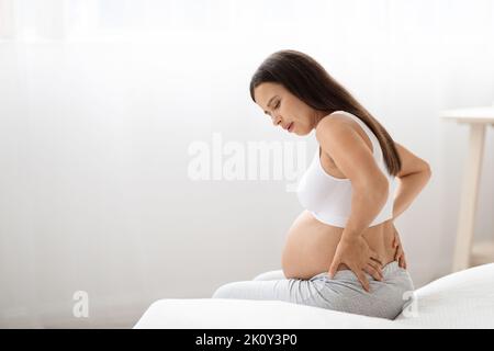 Expecting lady having back pain, sitting on bed at home Stock Photo