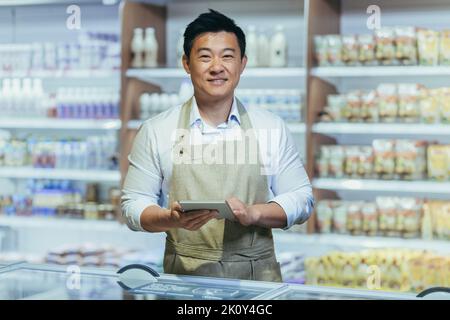 portrait of happy asian male small business owner standing in grocery store behind counter. Male manager with a tablet looking at the camera. friendly administrator food market. worker in apron. Stock Photo