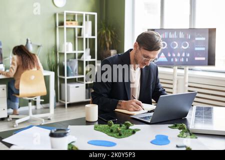 Young engineer making notes in notebook while working at his workplace with laptop at office Stock Photo