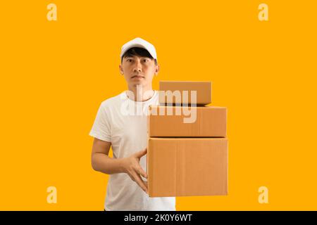 Young Asian delivery man wearing cap and white blank t-shirt holding parcel post box isolated on yellow background. express delivery service concept. Stock Photo