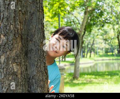 Pretty little girl peeking out from behind a tree. Portrait of child girl enjoying summer day in the green park. Stock Photo