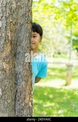 Pretty little girl peeking out from behind a tree. Portrait of child girl enjoying summer day in the green park. Stock Photo