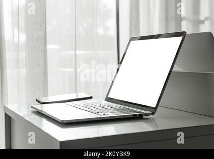 Computer laptop with blank screen and smartphone on white table in home interior or office background. Stock Photo