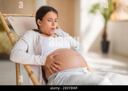 Pregnancy Pain. Pregnant Woman Touching Belly While Sitting In Chair At Home Stock Photo