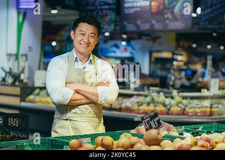 portrait of a happy handsome Asian salesman. Vegetables and fruits at the market or grocery store. Seller. a man in an apron looks at the camera and smiles farmer, small business owner greengrocer Stock Photo