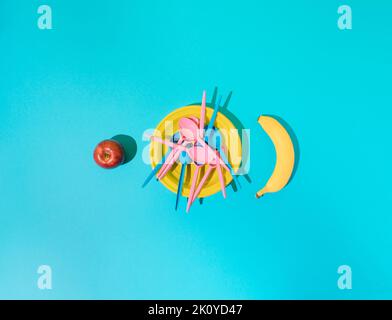 Yellow plastic plate with blue and pink plastic spoons and apple and banana on the sides on blue background. Minimal concept. Stock Photo