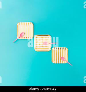Picnic setup with three gold and white striped paper plates and three plastic pink spoons on blue background. Minimal concept. Stock Photo