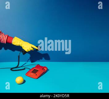 Minimal retro concept with hand in yellow cleaning glove answering red vintage phone on blue background. Stock Photo
