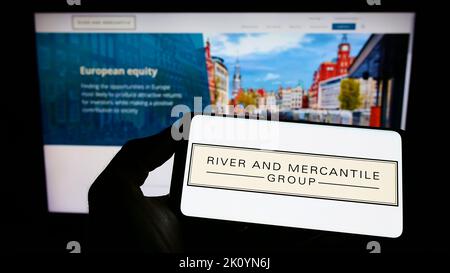 Person holding mobile phone with logo of company River and Mercantile Group Limited on screen in front of web page. Focus on phone display. Stock Photo