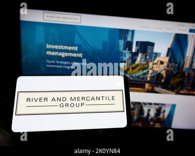 Person holding cellphone with logo of company River and Mercantile Group Limited on screen in front of business webpage. Focus on phone display. Stock Photo