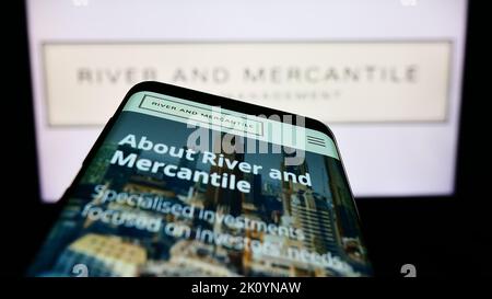 Smartphone with webpage of company River and Mercantile Group Limited on screen in front of business logo. Focus on top-left of phone display. Stock Photo